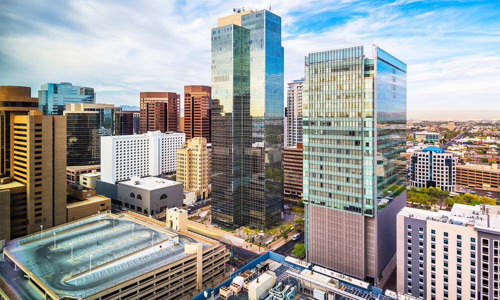 An aerial view of the modern downtown Phoenix skyline, showcasing tall glass skyscrapers and surrounding urban architecture under a bright blue sky.