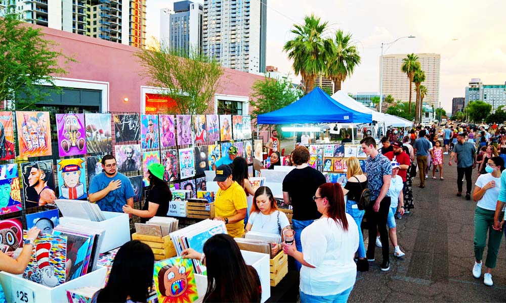 A vibrant outdoor art market at the First Fridays Art Walk in downtown Phoenix, Arizona, featuring colorful art displays, bustling crowds, and an urban cityscape in the background.